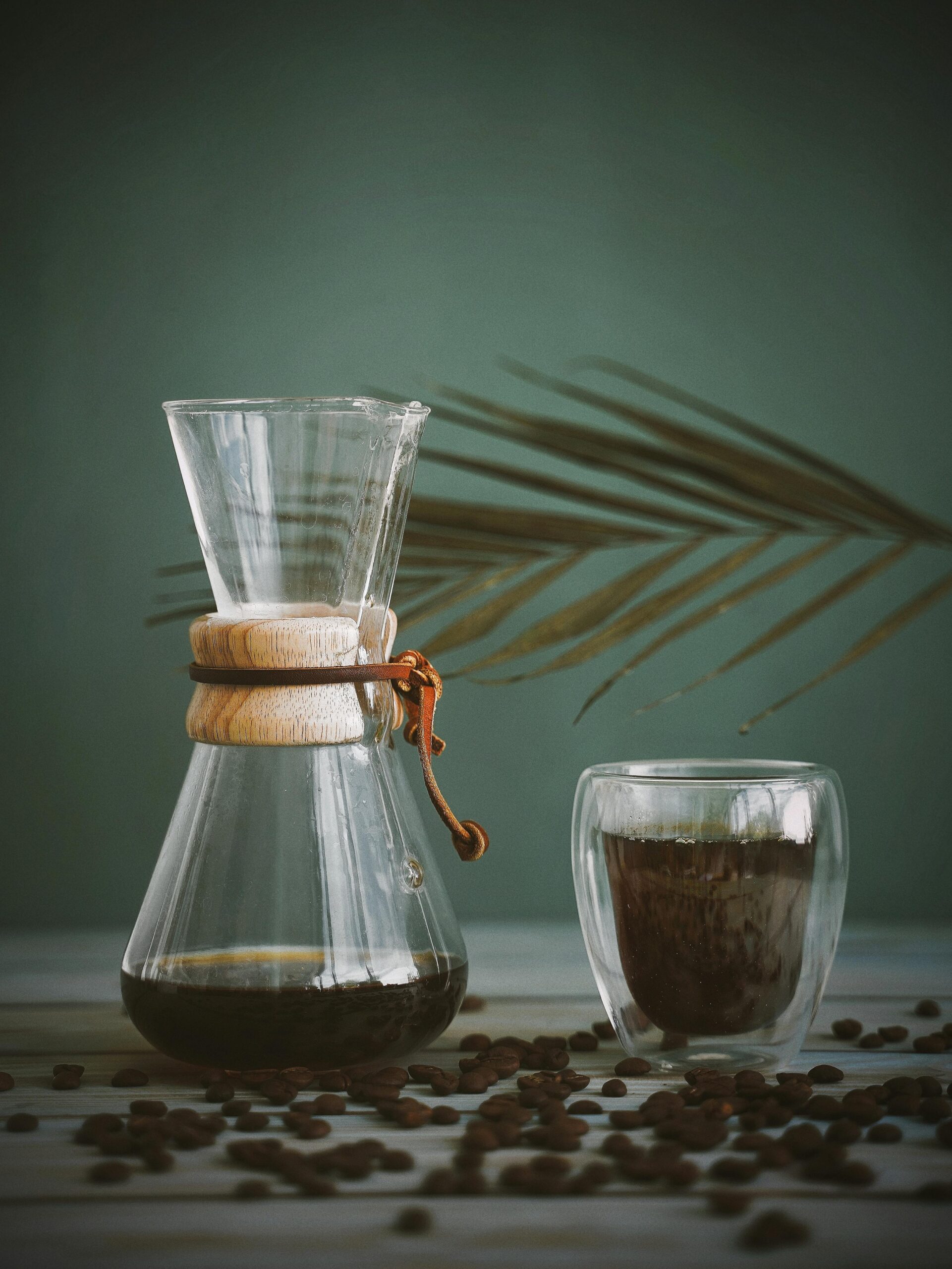 A clear glass coffee maker with a wooden collar and a small transparent double-walled glass cup, filled with coffee, placed on a table with coffee beans scattered around, with a green background