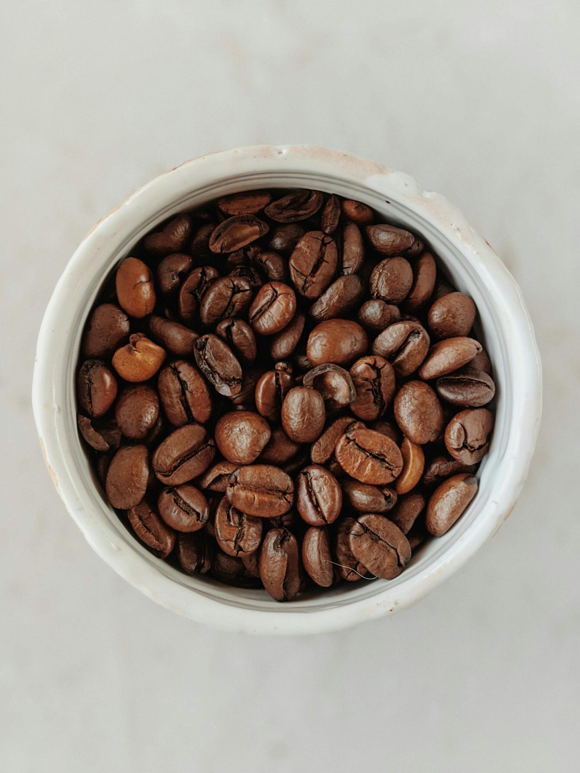A close-up overhead shot of roasted coffee beans in a white ceramic bowl on a light surface