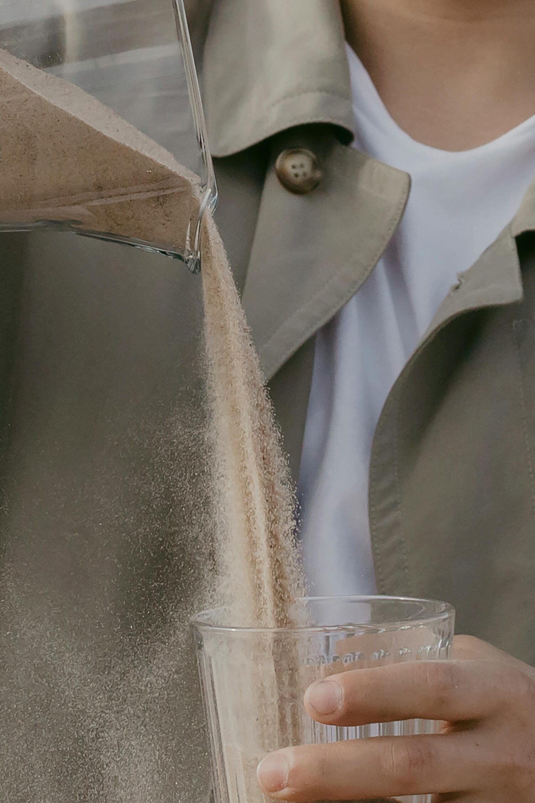A close-up shot of someone pouring a fine powder into a glass, with a soft-focus background and a warm-toned jacket visible, creating a natural and lifestyle vibe