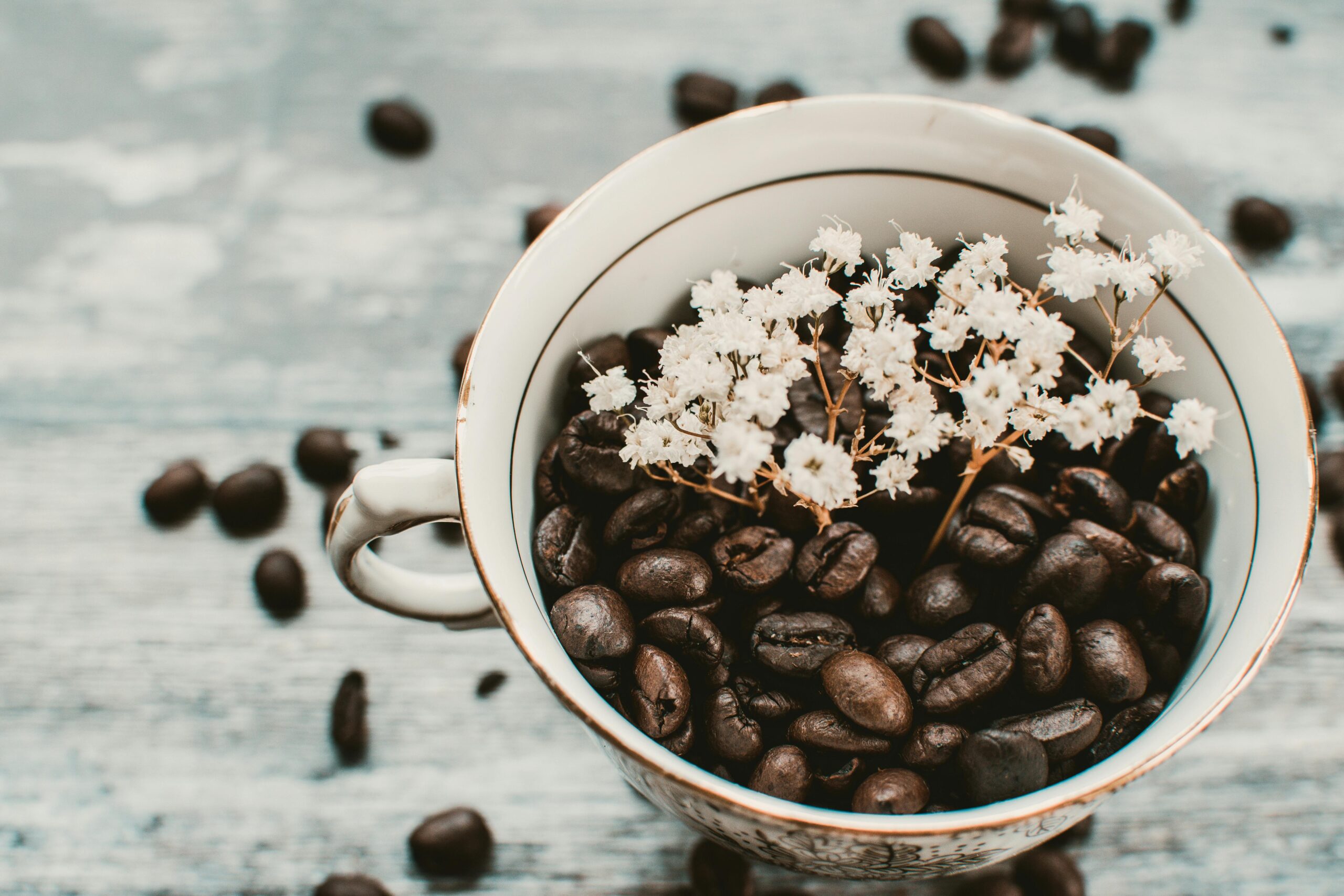 A vintage-style teacup filled with dark roasted coffee beans, adorned with small white flowers on top. Coffee beans are scattered on the wooden surface around the cup, enhancing the rustic charm