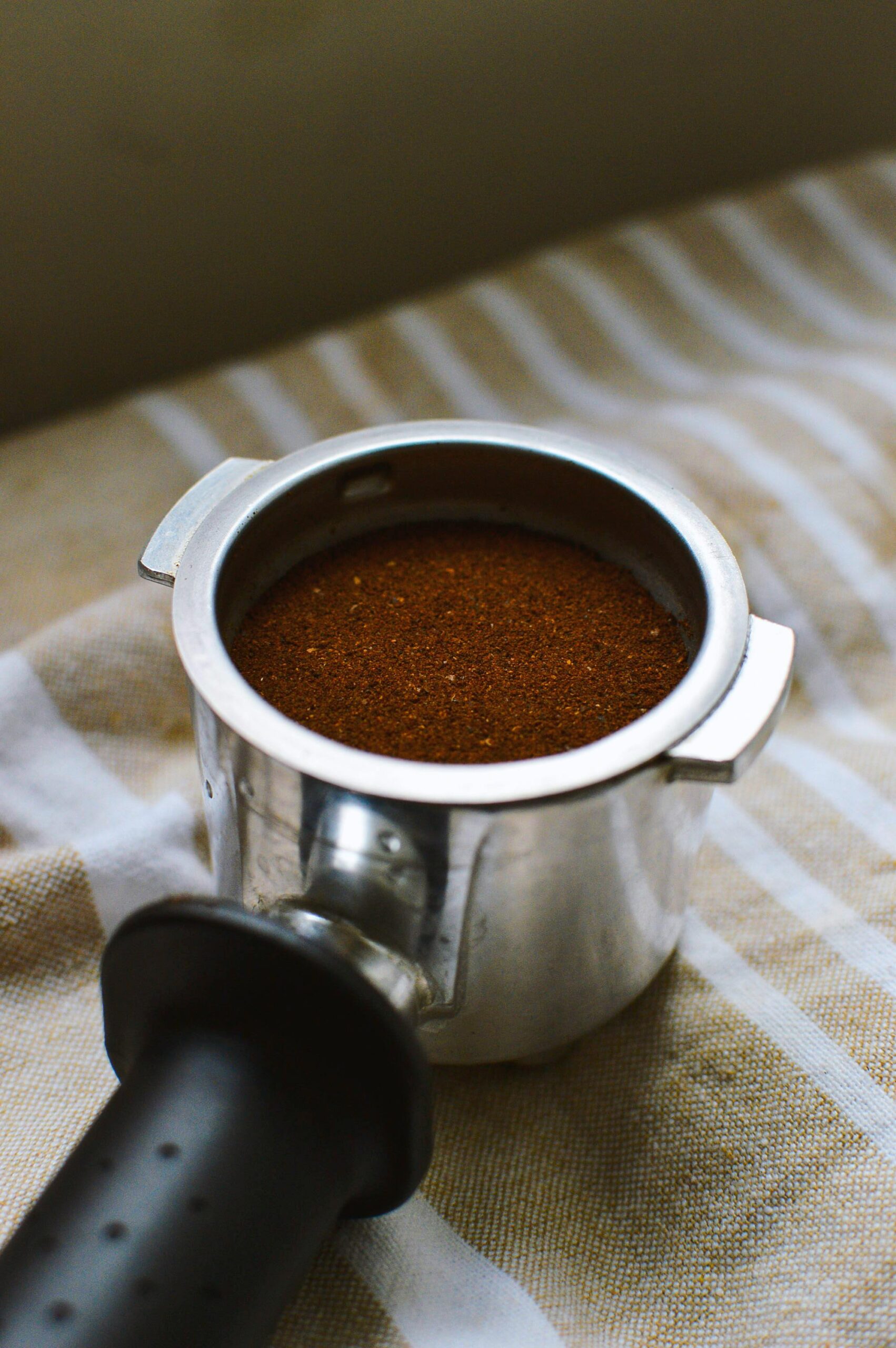 This image shows a portafilter filled with freshly ground coffee, ready for brewing. The finely ground coffee sits evenly in the portafilter, and the metal edges and sturdy handle hint at a well-crafted tool essential for making espresso. The background features a soft, striped fabric, adding a warm, cozy feel to the image, ideal for highlighting coffee preparation rituals