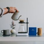 A person pouring hot water from a gooseneck kettle into a pour-over coffee maker. The coffee is dripping into a glass carafe positioned on a stand. Beside the setup is a gray coffee mug, a small plant, a blue coffee box, and a wooden coffee grinder on a modern, minimalist table