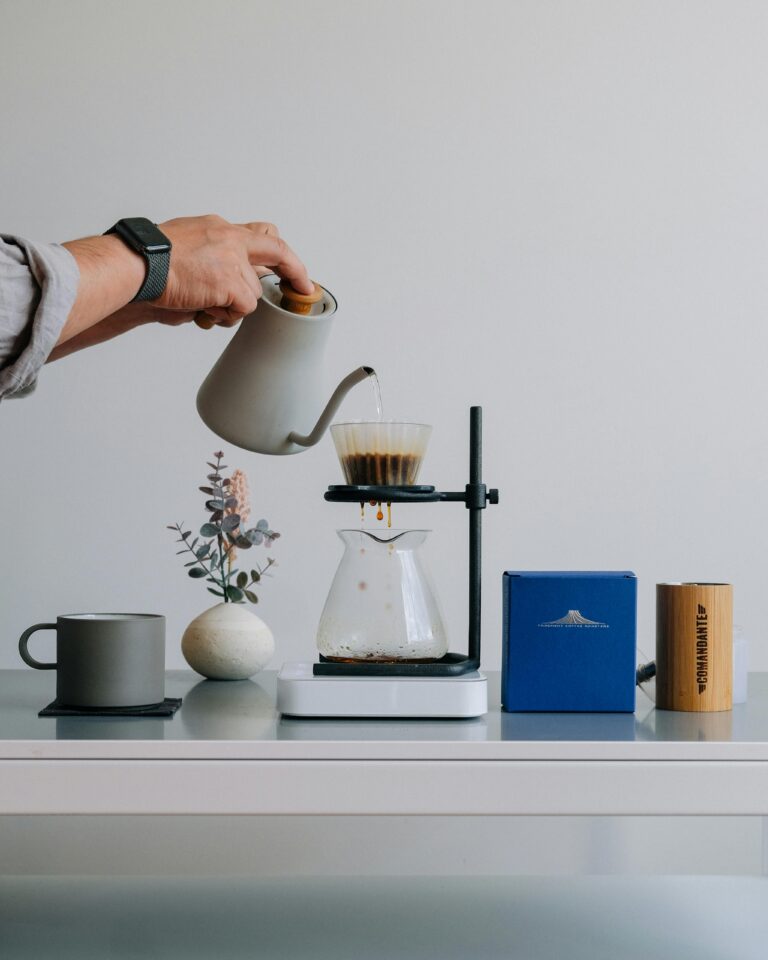 A person pouring hot water from a gooseneck kettle into a pour-over coffee maker. The coffee is dripping into a glass carafe positioned on a stand. Beside the setup is a gray coffee mug, a small plant, a blue coffee box, and a wooden coffee grinder on a modern, minimalist table