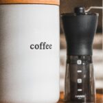 A ceramic container labeled "coffee" with a wooden lid, next to a manual coffee grinder. The container has a simple, minimalistic design, and the background is blurred, creating a cozy atmosphere