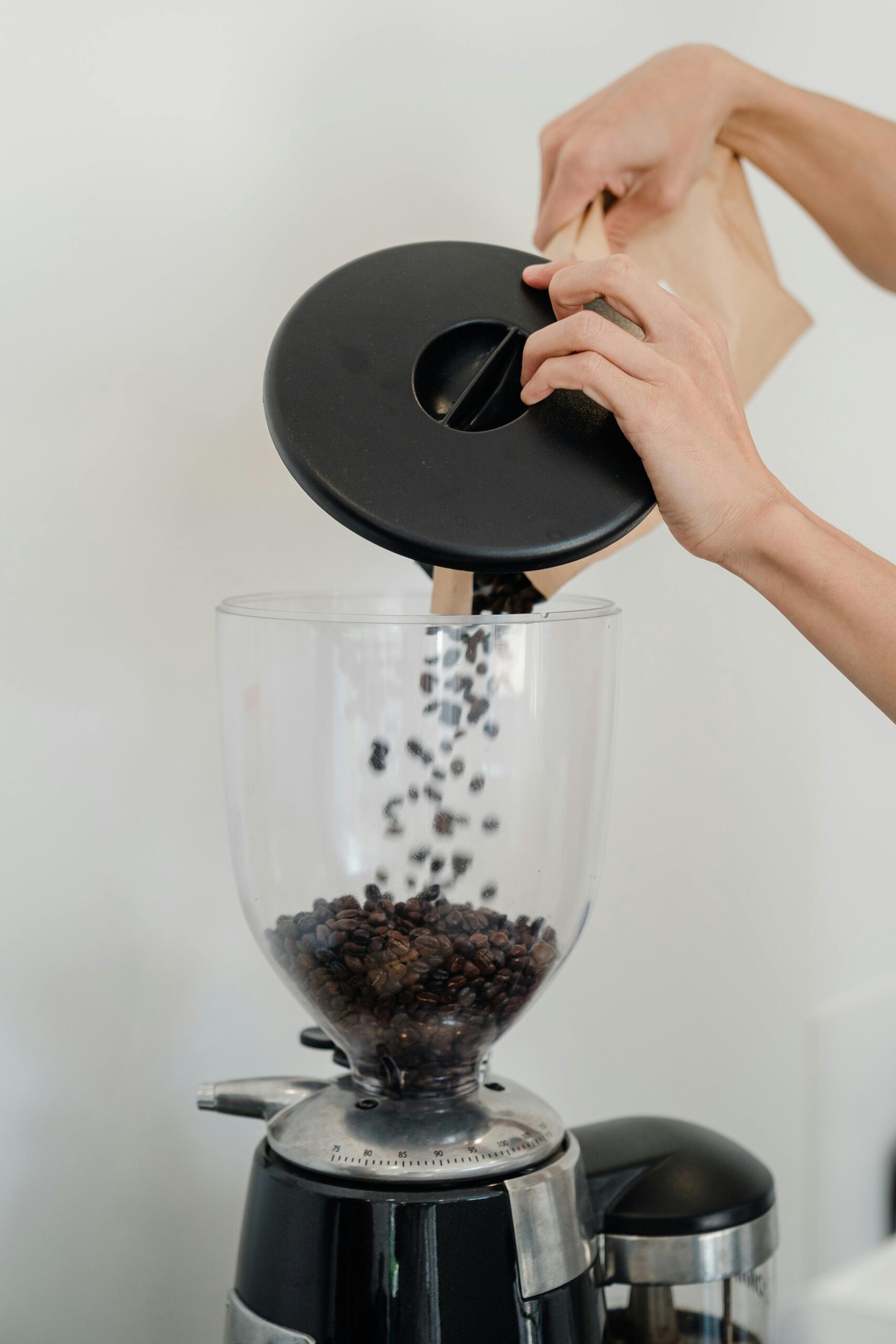 A close-up of a person pouring coffee beans from a brown kraft paper bag into a modern electric coffee grinder, set against a plain white background