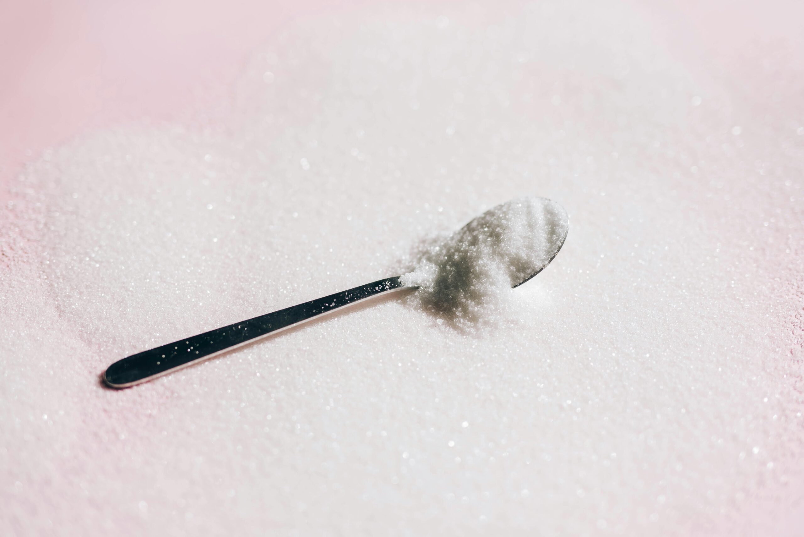 A spoon rests on a pink surface, surrounded by granulated white sugar, highlighting the texture and crystal-like appearance of the sugar