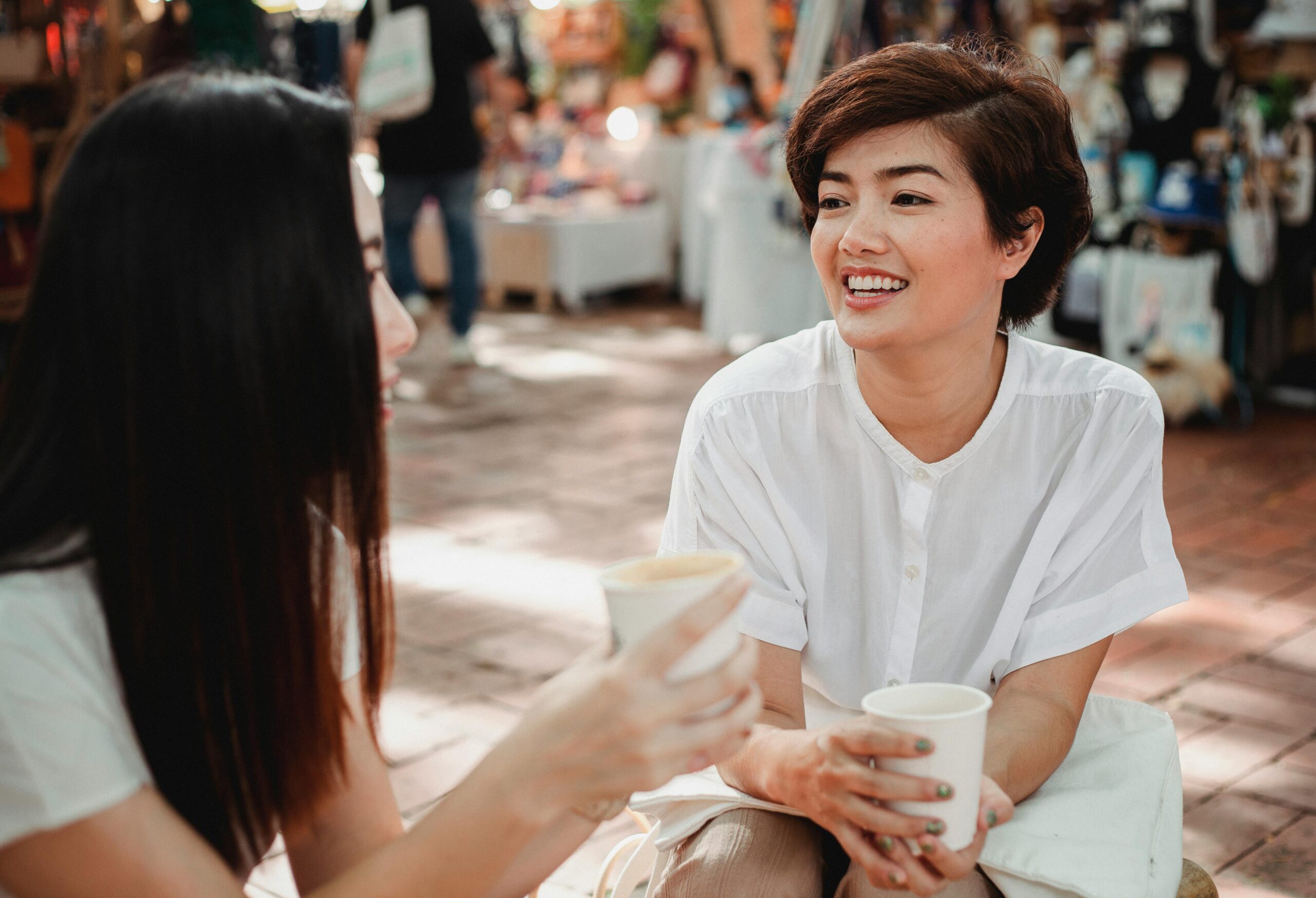The image shows two women sitting outdoors, enjoying a casual conversation while holding takeaway cups, presumably filled with coffee. One woman, wearing a white shirt, has short hair and is smiling warmly. The background suggests a market or outdoor gathering, with blurred figures and items in stalls behind them, creating a relaxed and friendly atmosphere