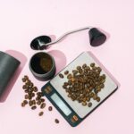 Top-down view of coffee brewing essentials on a pink background, featuring whole coffee beans on a digital scale, a manual coffee grinder with freshly ground coffee inside, and a grinder handle placed nearby. The setup highlights the tools needed to prepare freshly ground coffee