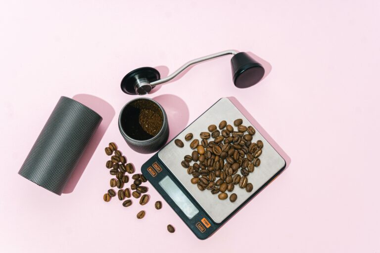 Top-down view of coffee brewing essentials on a pink background, featuring whole coffee beans on a digital scale, a manual coffee grinder with freshly ground coffee inside, and a grinder handle placed nearby. The setup highlights the tools needed to prepare freshly ground coffee