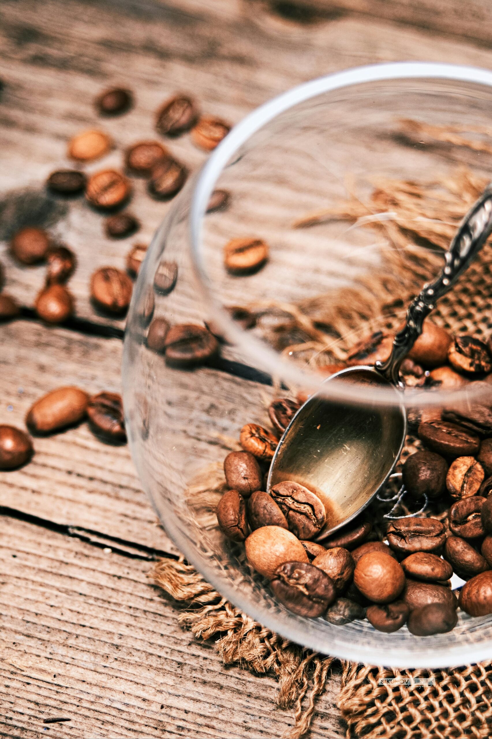 A metal spoon in a clear glass container scooping coffee beans, with additional beans scattered across a rustic wooden surface