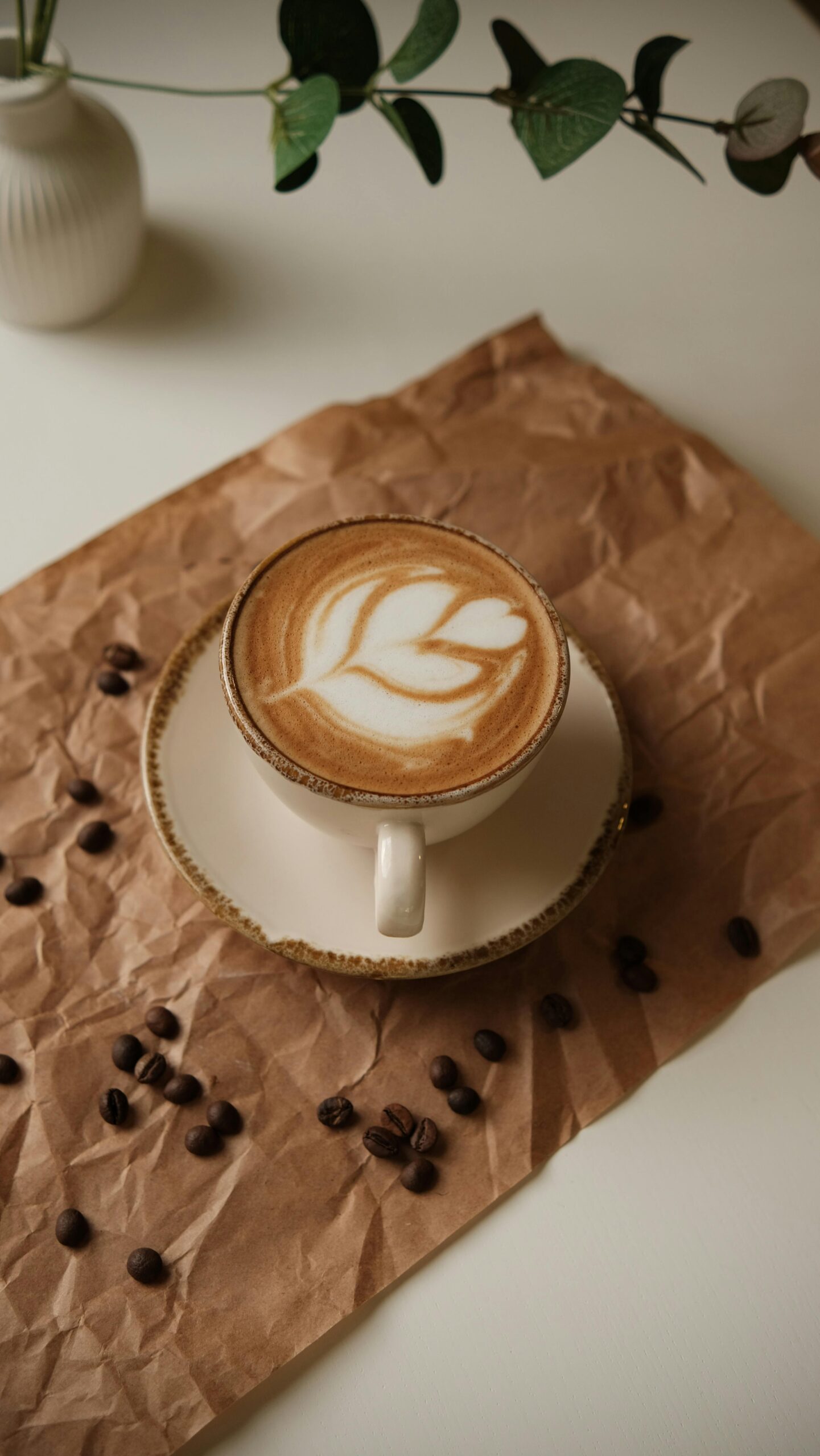 A cup of latte art on a white saucer, placed on crumpled brown paper with coffee beans scattered around, and a small vase with green leaves in the background