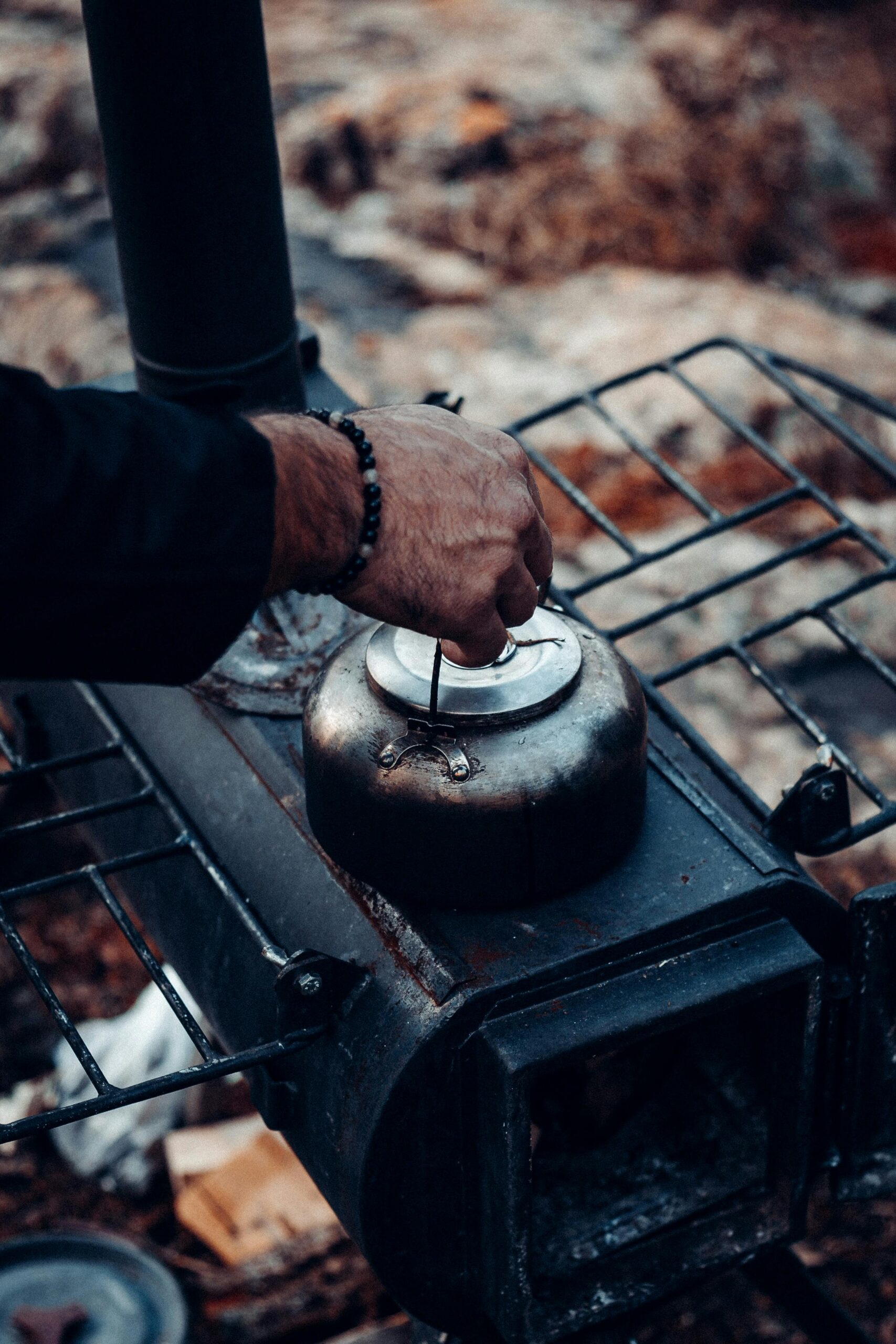A close-up of a person's hand lifting the lid of a rustic metal kettle placed on an outdoor wood stove, surrounded by a rugged natural environment