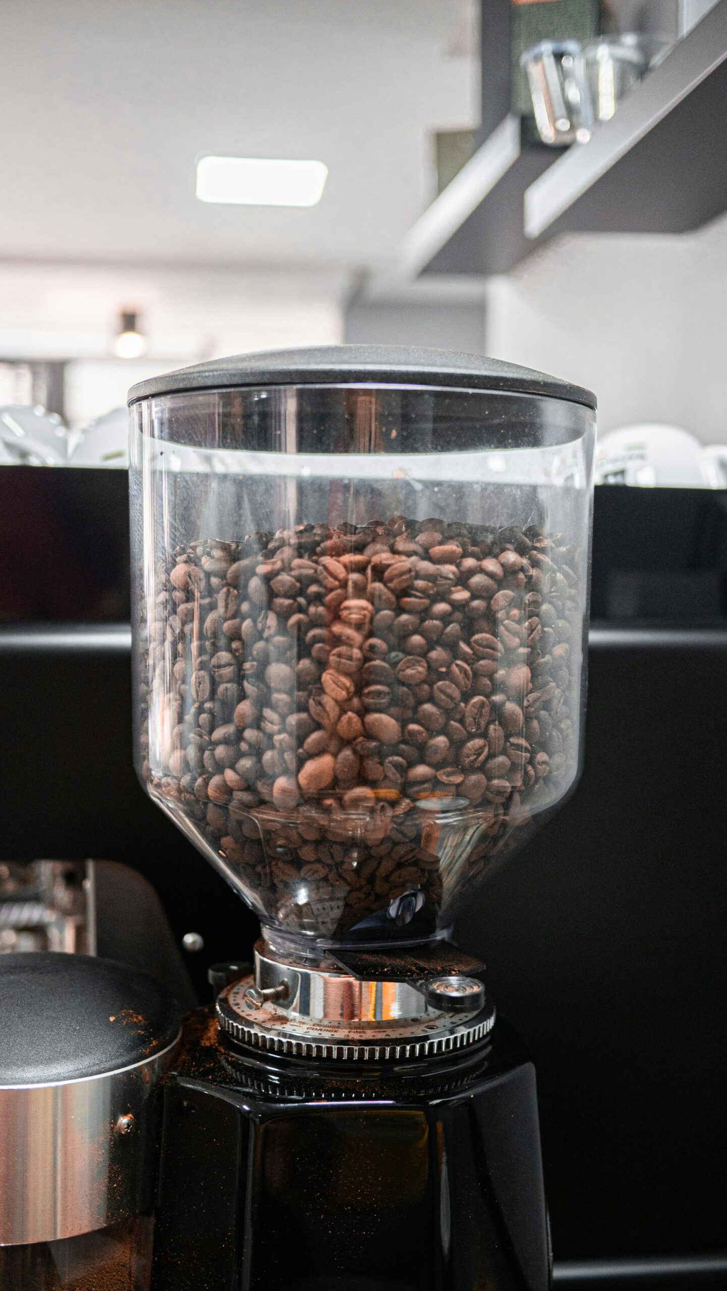 A close-up of a coffee grinder filled with roasted coffee beans in a transparent hopper, set in a modern café environment