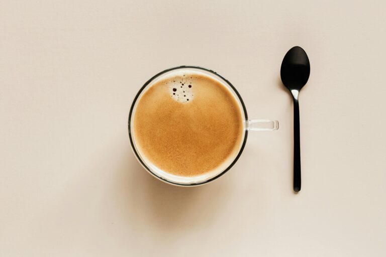 A top-down view of a glass cup filled with freshly brewed coffee, featuring a smooth crema layer on top. A black spoon is placed beside the cup on a clean, neutral-toned background