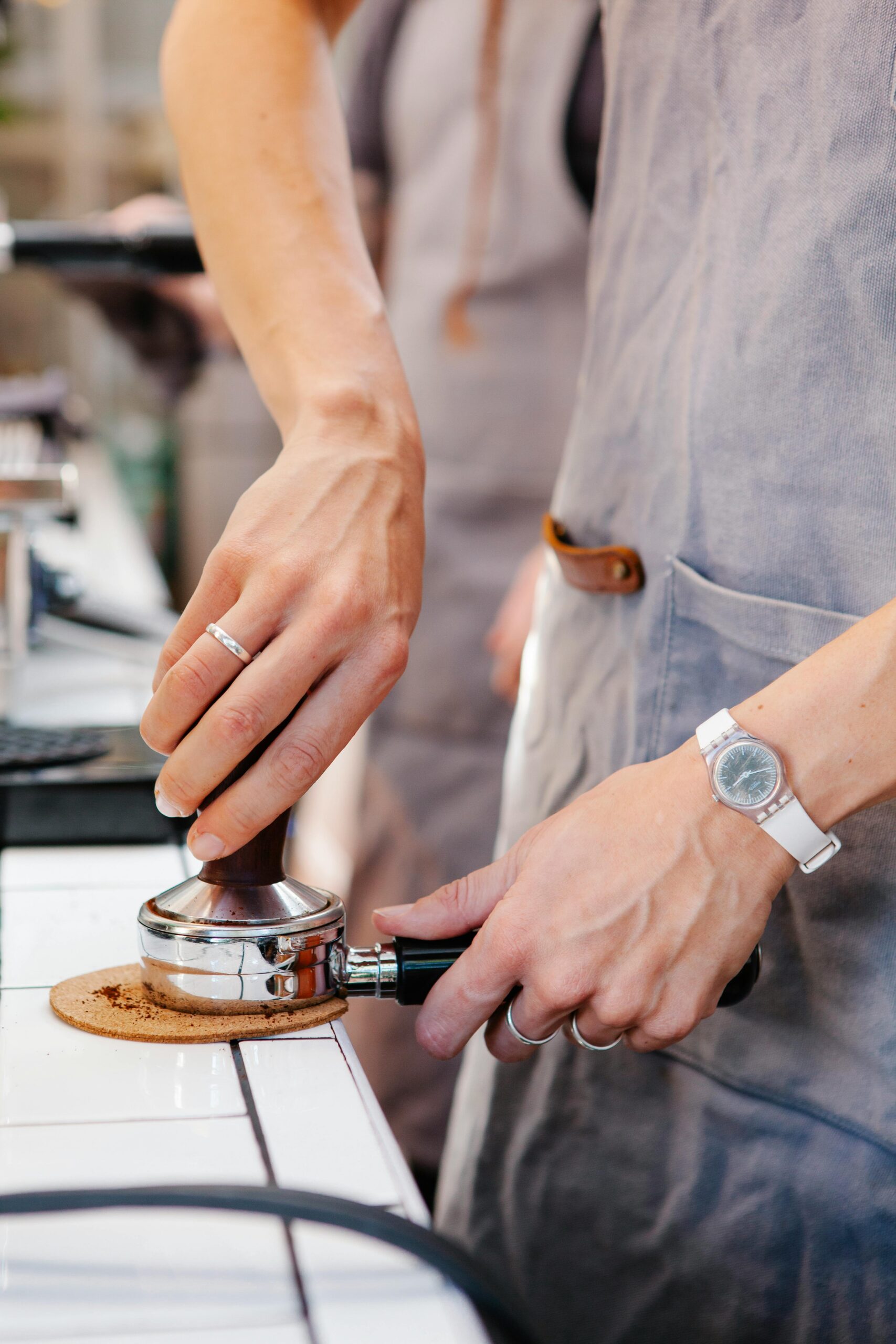 A barista wearing a gray apron firmly tamping freshly ground coffee into a portafilter, preparing for espresso extraction in a café setting